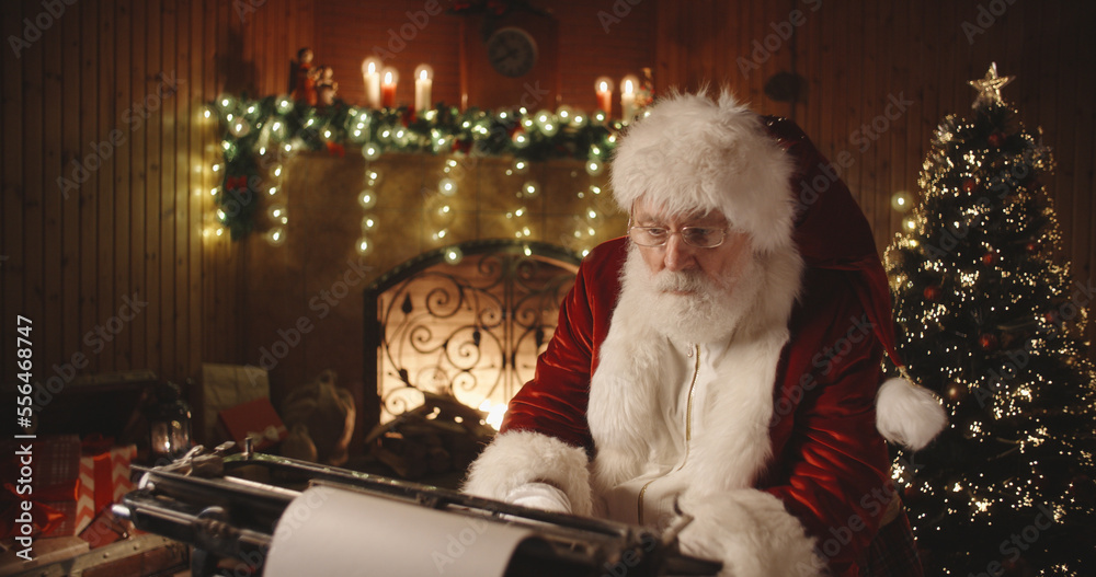 Senior bearded man in santa claus clothing is working on letter at his typewriter then looking at camera - christmas spirit, holidays and celebrations concept 