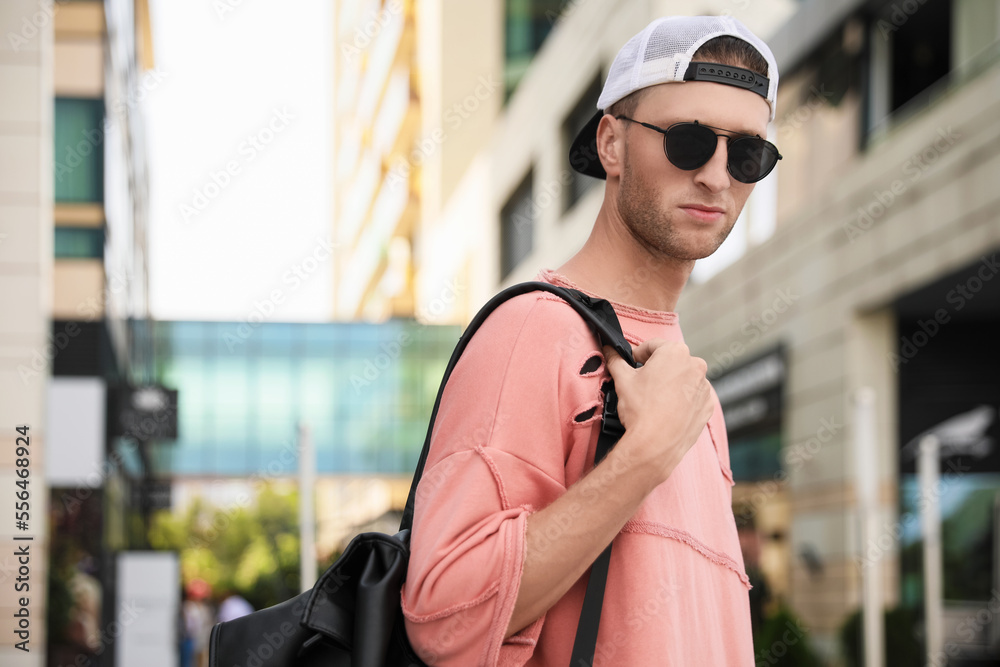 Handsome young man with stylish sunglasses and backpack on city street, space for text