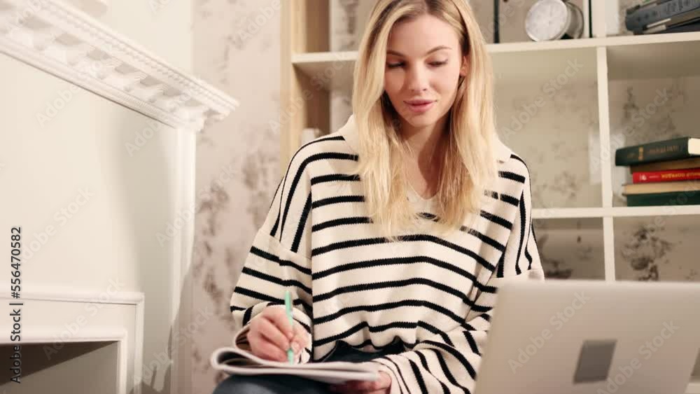 Attractive young blond girl preparing for exam and studying at home Charming young student focused on learning and make notes in her notebook while looking at laptop computer screen indoors Attractive young blond girl preparing for exam and studying at home Charming young student focused on learning and make notes in her notebook while looking at laptop computer screen indoors