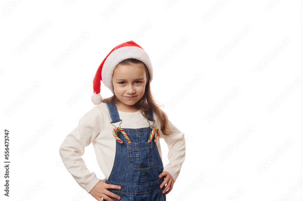 Adorable Caucasian little child girl wearing Santa hat, posing with hands on waist, with Christmas striped candy canes on the pocket of his denim overalls, looking at camera, against white background Adorable Caucasian little child girl wearing Santa hat, posing with hands on waist, with Christmas striped candy canes on the pocket of his denim overalls, looking at camera, against white background