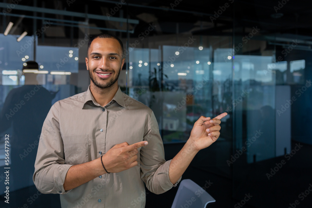 Obraz premium Hispanic businessman inside the office smiling and looking at the camera while standing and pointing to the side with his hands, a man in a casual shirt near the window.