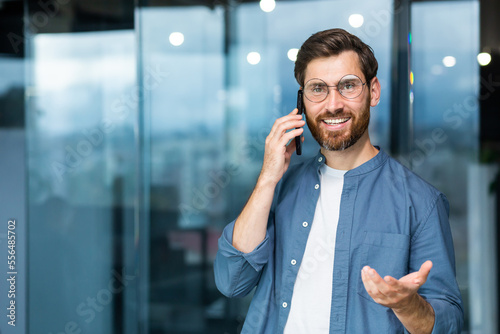 Portrait of a mature successful businessman inside the office, the man is smiling and looking at the camera by the window while standing and talking cheerfully and smiling on the phone.