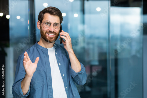 Portrait of a mature successful businessman inside the office, the man is smiling and looking at the camera by the window while standing and talking cheerfully and smiling on the phone.