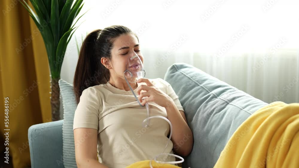 Beautiful young woman with brown hair doing inhalation at home, inhaling through inhaler mask, looking happy and satisfied with the treatment, smiling at the end. Beautiful young woman with brown hair doing inhalation at home, inhaling through inhaler mask, looking happy and satisfied with the treatment, smiling at the end.