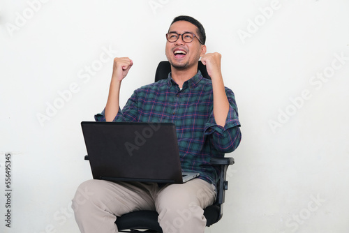 Asian man sitting in an office chair with laptop on his thigh showing excitement photo