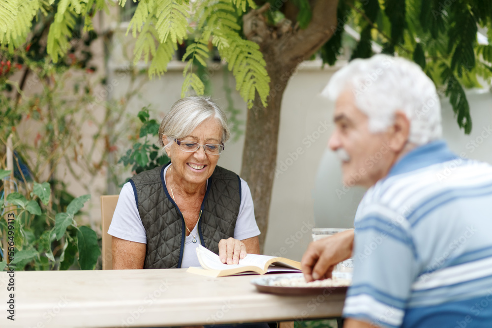 Loving elderly spouses resting in garden and enjoying days, sitting outdoors.Woman reading book, man in front of her Loving elderly spouses resting in garden and enjoying days, sitting outdoors.Woman reading book, man in front of her