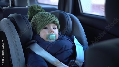 A little boy is sitting in a child seat in a car. A baby travels in a car in winter. Boy in jacket and hat.