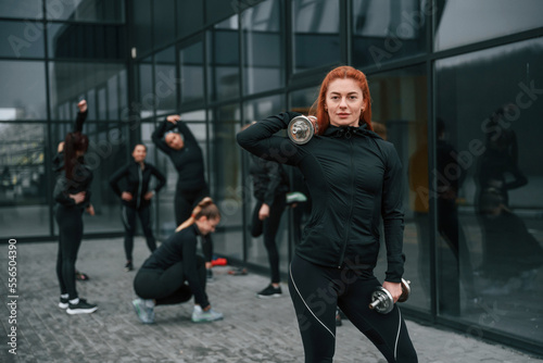 Lifting dumbbells. Group of sportive women is outdoors near black building