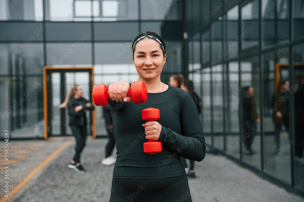 Smiling, doing exercises by using dumbbells. Group of sportive women is outdoors near black building