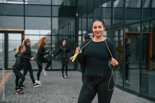 With yellow colored jump rope. Group of sportive women is outdoors near black building