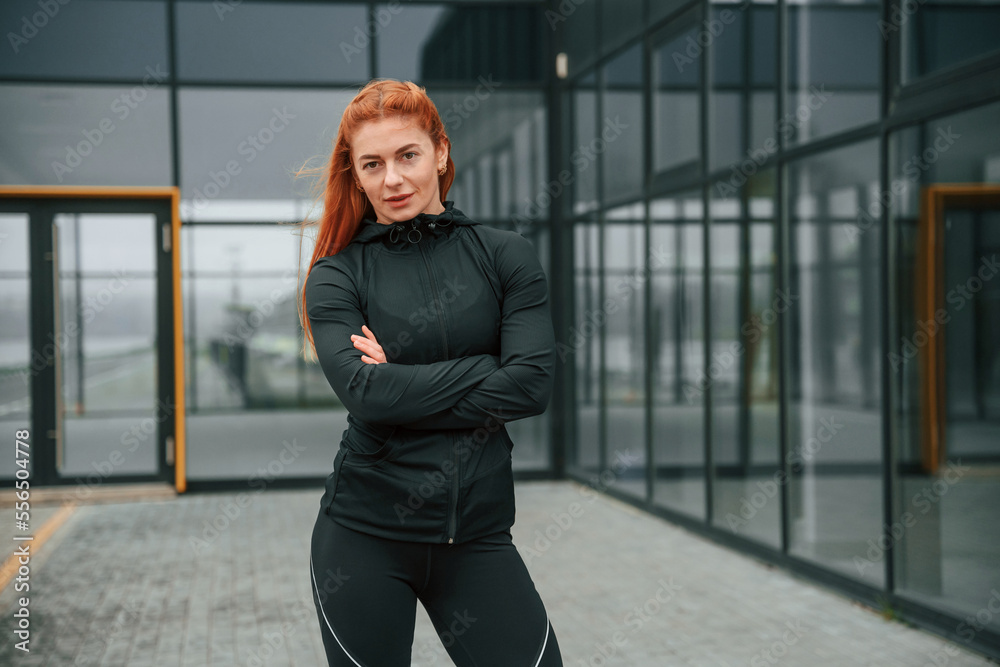 Obraz premium Standing with arms crossed. Woman is sportive clothes is outdoors against black building