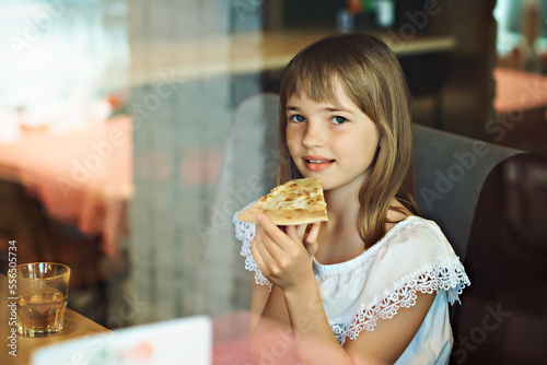 A young girl with long hair is eating a slice of pizza. The photo was taken through the window of the pizzeria.