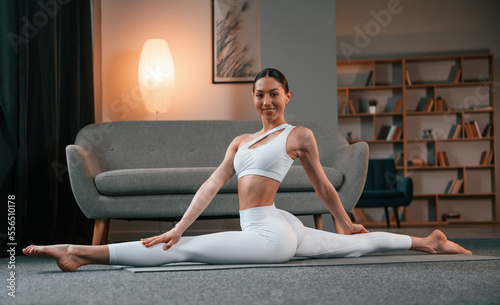 Practicing on the floor. Young woman with slim body type in white sportswear is indoors doing yoga