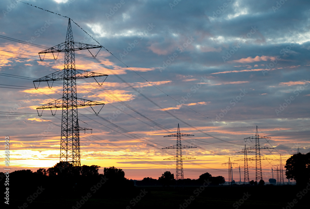 Strommasten mit Stromkabeln vor der untergehenden Sonne mit Wolken, Farbenspiel.