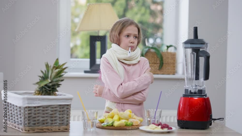 Sad ill Caucasian girl with thermometer in mouth standing at table with healthful fruits measuring body temperature. Medium shot portrait of upset unwell teenager at home with symptoms of illness
