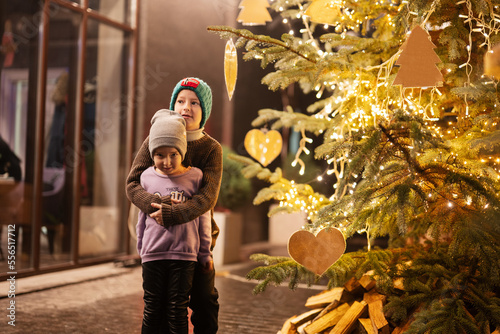Brother with sister having fun near illuminated Christmas tree outdoor in evening. photo