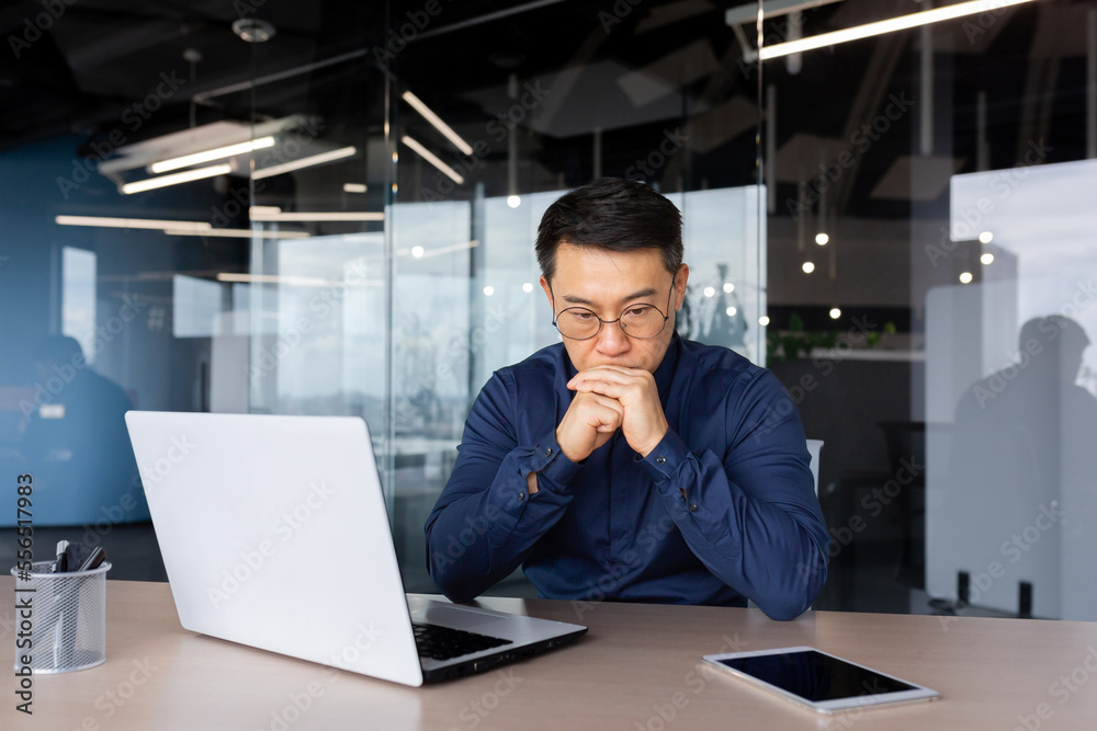 Serious male Asian businessman sits in the office at the table, works with a laptop and documents. He holds his head in his hands, thinks, solves a problem. Serious male Asian businessman sits in the office at the table, works with a laptop and documents. He holds his head in his hands, thinks, solves a problem.