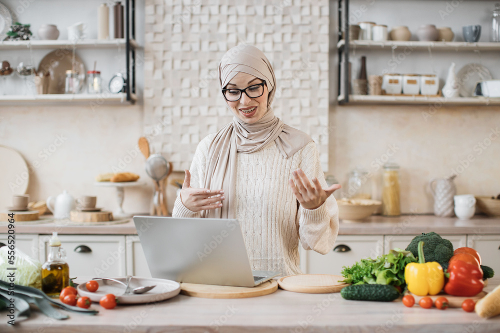 Muslim woman cooking delicious and healthy food in modern kitchen looking at laptop, while having online class, lesson or having video call with followers in social networks, media in internet at home Muslim woman cooking delicious and healthy food in modern kitchen looking at laptop, while having online class, lesson or having video call with followers in social networks, media in internet at home