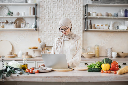 Vegetarian food, healthy eating, people, technology and diet concept. Attractive muslim woman preparing vegetable salad for breakfast standing near table looking on laptop pc computer at home kitchen.