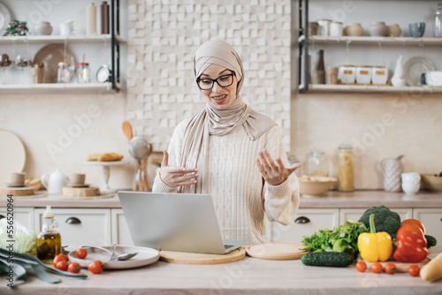 Muslim woman cooking delicious and healthy food in modern kitchen looking at laptop, while having online class, lesson or having video call with followers in social networks, media in internet at home