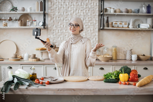 Muslim female having video call using selfie stick while providing cooking online class healthy vegan food Happy young woman blogger showing to her followers how to prepare dinner with fresh vegetable