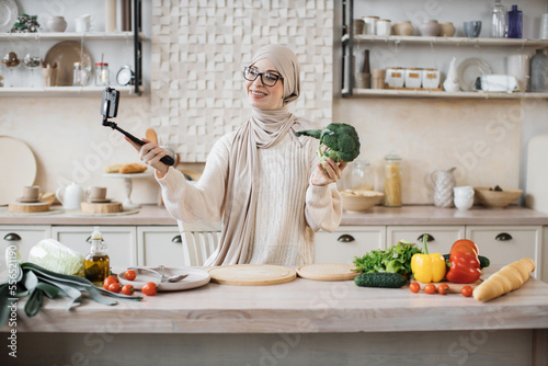 Happy young muslim woman using smartphone in selfie stick standing in kitchen in the morning Happy female blogger holding broccoli speaking with followers in social media about preparing healthy salad