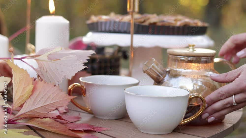 Cozy autumn picnic in the park. Close up of unrecognisable woman pouring hot tea from glass kettle into classic cups. Homemade apple pie, maple leaves and burning candles on wooden table outdoors