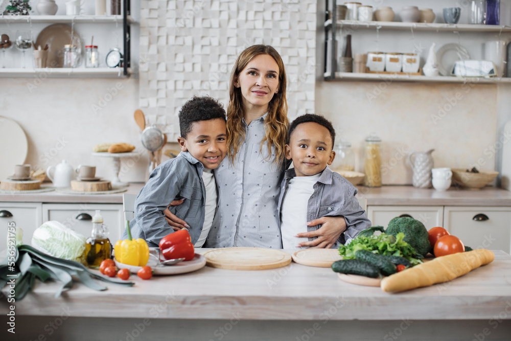 Healthy food, home resting concept. Pretty young mother and two children sons preparing salad with fresh vegetables, hugging and smiling looking at the camera while cooking in kitchen. Healthy food, home resting concept. Pretty young mother and two children sons preparing salad with fresh vegetables, hugging and smiling looking at the camera while cooking in kitchen.