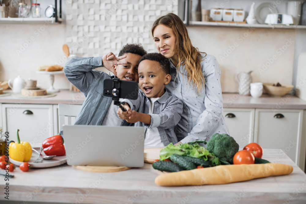 Pretty young mother and two children sons preparing salad with fresh vegetables, hugging and smiling, doing selfie using smart phone and stick while cooking in kitchen.