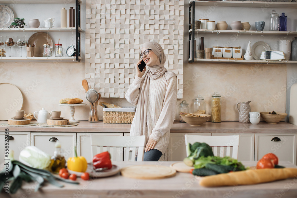 Portrait of young beautiful muslim woman in hijab standing at bright modern kitchen, using phone during call with her friends while cooking vegan salad from fresh vegetables at home. Portrait of young beautiful muslim woman in hijab standing at bright modern kitchen, using phone during call with her friends while cooking vegan salad from fresh vegetables at home.