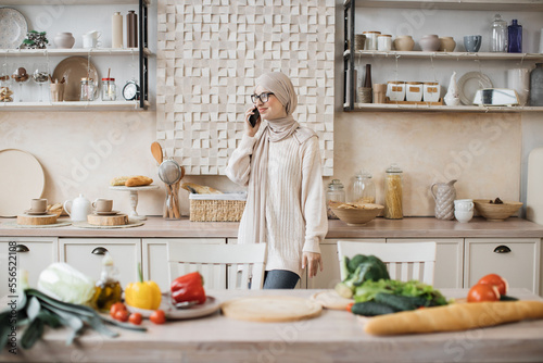 Portrait of young beautiful muslim woman in hijab standing at bright modern kitchen, using phone during call with her friends while cooking vegan salad from fresh vegetables at home.