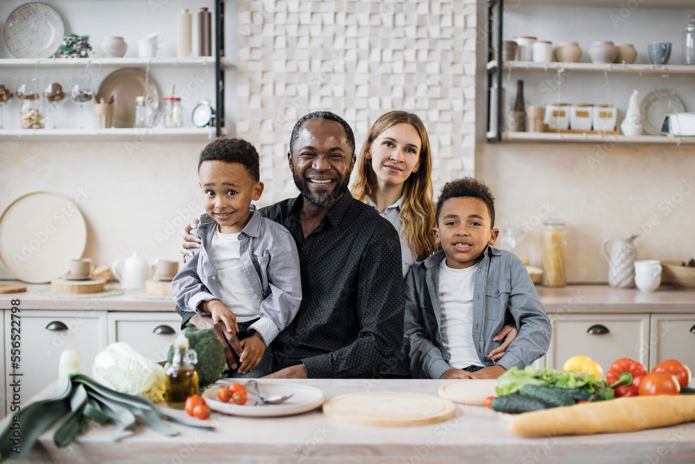 Healthy food, home resting concept. Pretty young mother, father and two children sons preparing salad with fresh vegetables, hugging and smiling looking at the camera while cooking in kitchen.