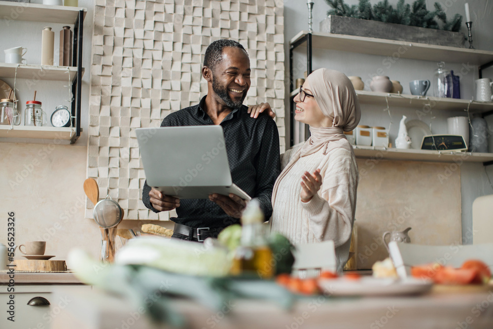 Young wife and husband looking at online recipe on a digital laptop pc. Attractive muslim female and african male surfing the internet for recipes while making vegetarian salad in bright kitchen. Young wife and husband looking at online recipe on a digital laptop pc. Attractive muslim female and african male surfing the internet for recipes while making vegetarian salad in bright kitchen.