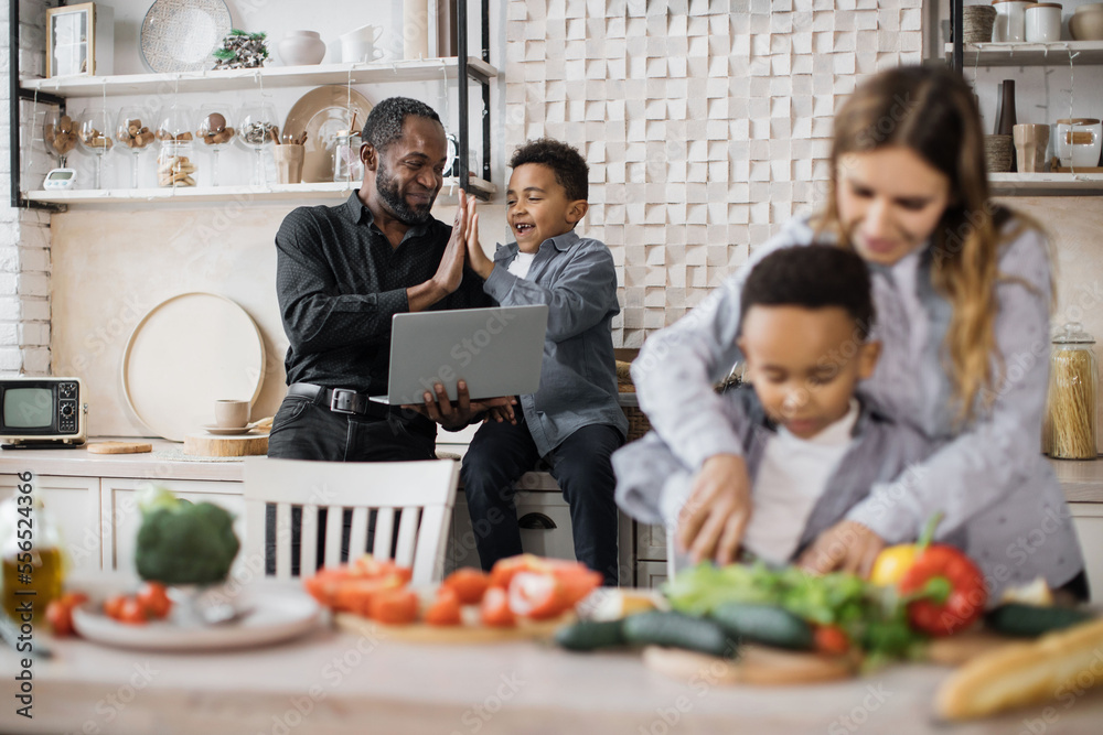 Focus on loving african father and small boy child finding recipe of holiday meal and giving high five. Happy mother teach little preschooler son chop vegetables preparing salad for lunch together.