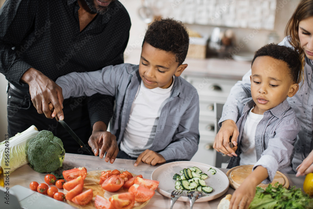 Close up view of happy multinational parents and two preschool sons preparing tasty food at kitchen. Mother and father teaching lovely kids to cook chopping fresh vegetables with knife.