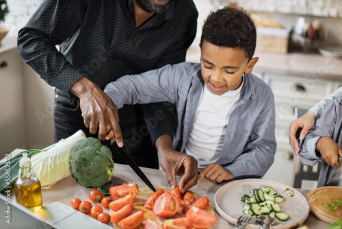 Cropped high angle view of cute african preschool boy helping his father to chop fresh vegetables tomatoes and cucumbers for delicious healthy salad. Little helper concept.