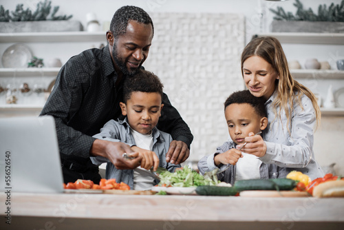 Young parents teaching african little cute sons preparing healthy vegetarian meal with sliced vegetables. Mom and dad sharing salad recipe  multiracial family chatting enjoying time in kitchen.