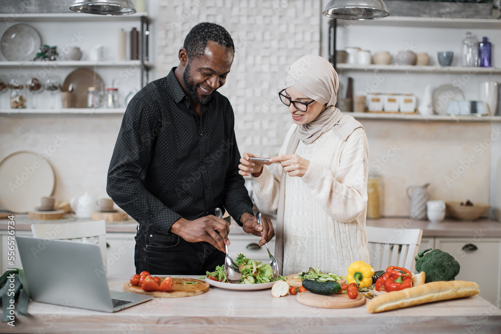 African american man holding spoon and fork, mixing ingredients in bowl, preparing delicious healthy salad, meal on table dinner in kitchen, next to his wife taking photos for social media. African american man holding spoon and fork, mixing ingredients in bowl, preparing delicious healthy salad, meal on table dinner in kitchen, next to his wife taking photos for social media.