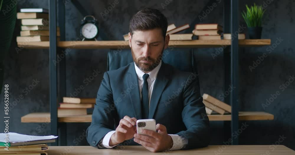 Serious bearded employee is writing a text message using a modern smartphone. Confident office worker in suit using smartphone while working in office. Millennial Employee Uses Mobile App at Work