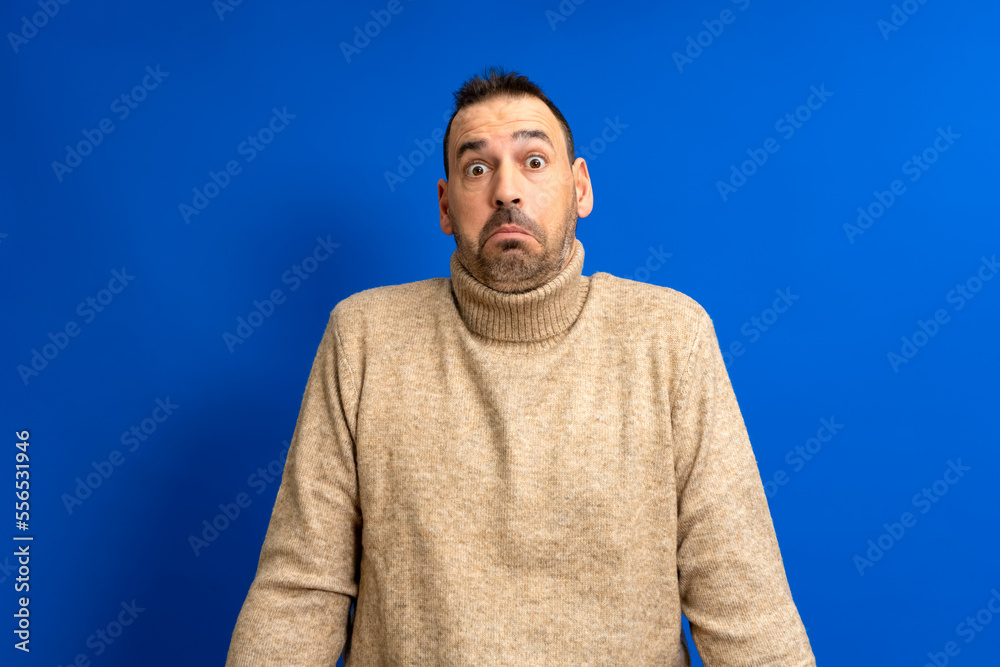 Fototapeta premium Frontal portrait of a hispanic man with a beard wearing a brown turtleneck doubting shrugging isolated on a blue background.