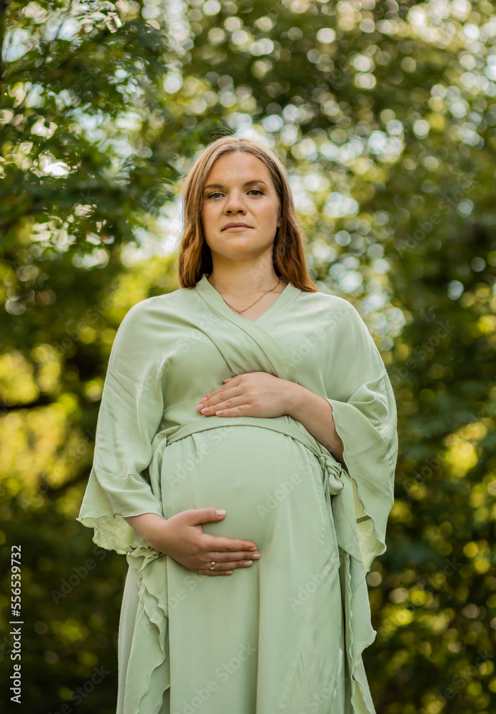 Young pregnant woman puts hands on big belly, green trees,blue sky on background. Female wears dress. Magic happy pregnancy. Child delivery preparation,emotional connection with baby.Vertical