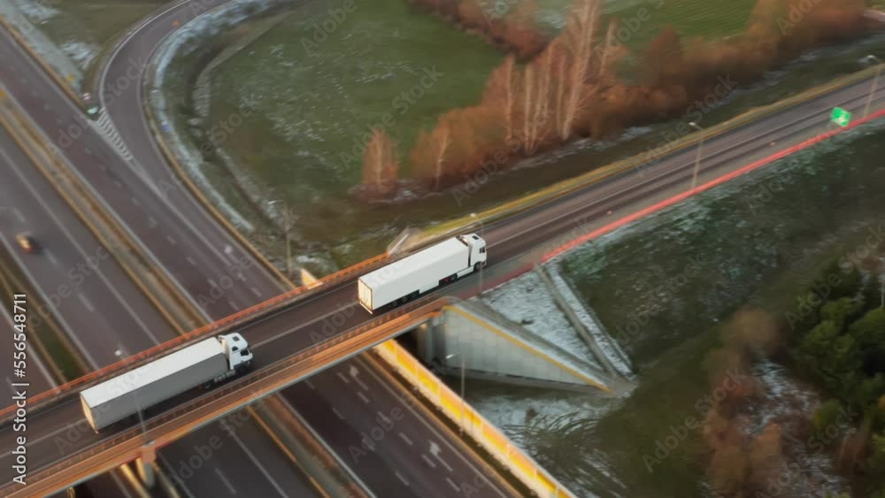A white truck with a trailer drives over a bridge crossing an expressway with heavy traffic of cars and trucks. Transportation and delivery of goods along the international cargo logistics route.