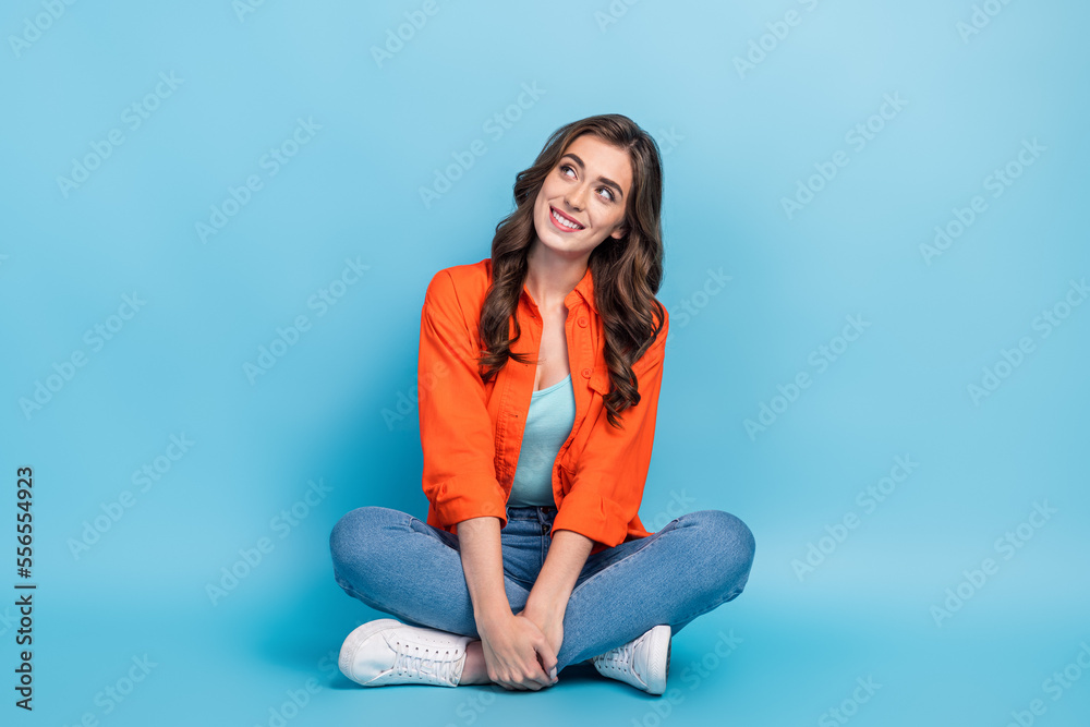 Full length photo of young attractive woman sitting floor look empty space wear trendy orange clothes isolated on blue color background Full length photo of young attractive woman sitting floor look empty space wear trendy orange clothes isolated on blue color background