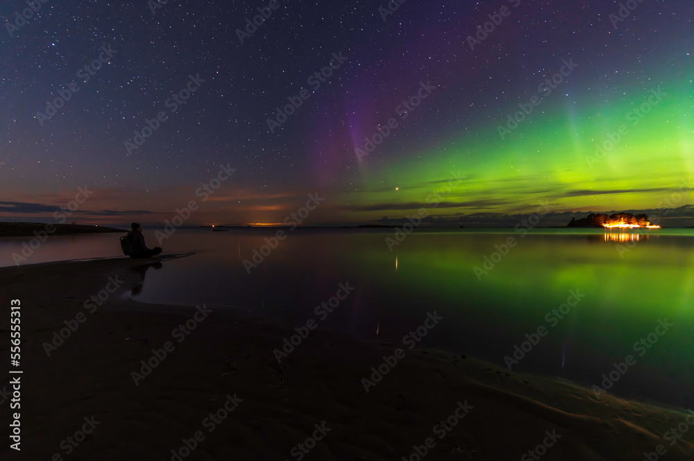 A man sitting on the beach looking at the northern lights. Reflections in the water. 
Storsand Jakobstad/Pietarsaari. Finland