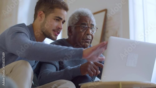 Bearded Caucasian guy helping senior Black man with using laptop. Young man sitting on couch with his grandpa while explaining things and pointing at screen. Family bonding, generations concept.