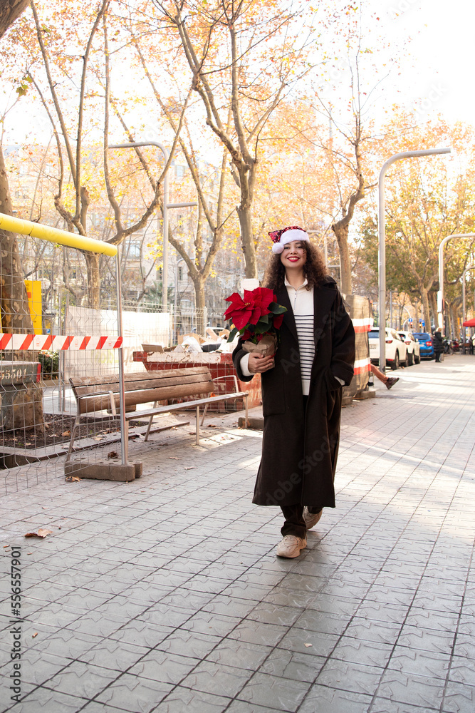 Christmas mood, full-length model in a coat and santa hat walks down the street with a flower in her hands