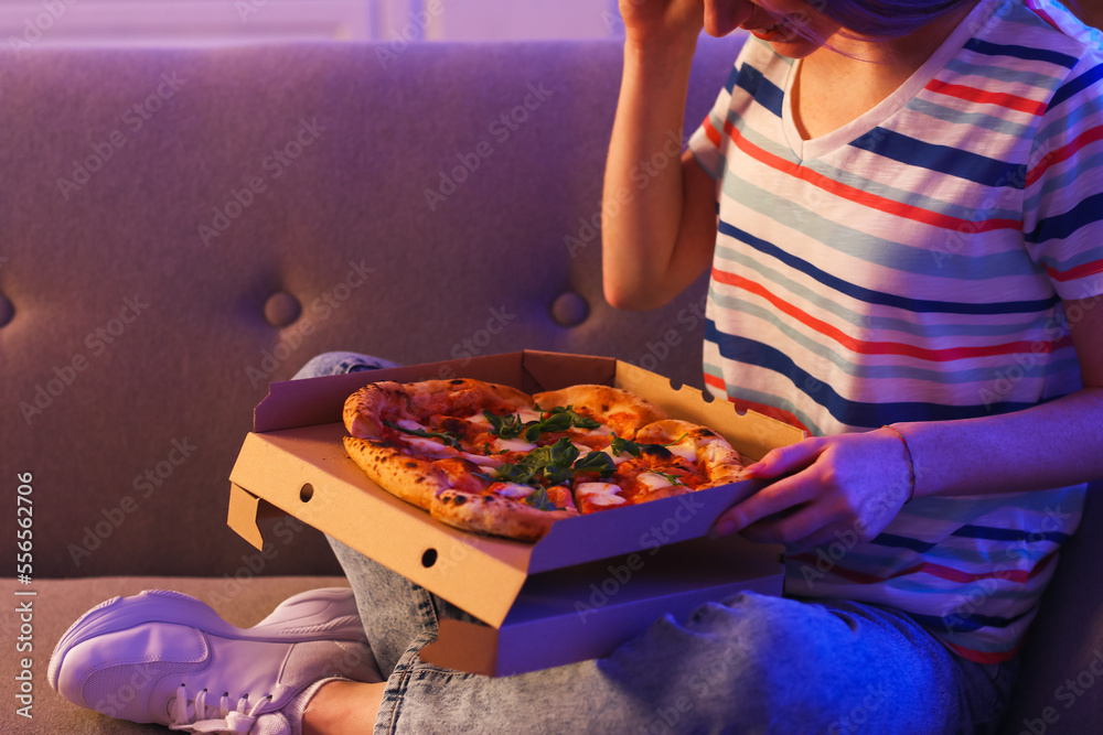 Young woman with tasty pizza sitting on sofa in kitchen at night