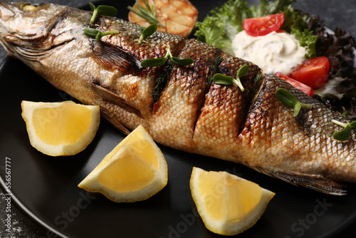 Delicious baked sea bass fish and ingredients on black plate, closeup