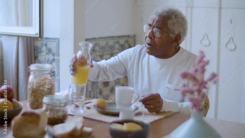 Black elderly man enjoying breakfast at home. Grey-haired senior man in glasses pouring and drinking orange juice and having cupcakes, cornflakes and fruit on table. Lifestyle, healthy food concept. Black elderly man enjoying breakfast at home. Grey-haired senior man in glasses pouring and drinking orange juice and having cupcakes, cornflakes and fruit on table. Lifestyle, healthy food concept.