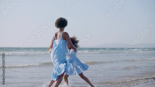 Cute Black girl and her beautiful mum having fun at seashore, wearing blue dresses. Young barefoot woman dancing and spinning, holding hands with daughter on the beach. Happy family, holiday concept.